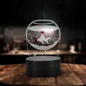 May include: A decorative night light with a fishbowl design, featuring a goldfish silhouette and bubbles. The lamp has a black base and a clear acrylic top with the text "Your Text". It is displayed on a wooden surface.