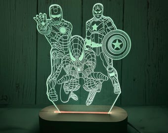 Könnte beinhalten: Ein leuchtendes LED-Nachtlicht mit Iron Man, Spider-Man und Captain America. Die Superheldenfiguren sind in einem detaillierten Strichstil dargestellt, mit einem Holzsockel und einem sanften, grünen Leuchten. Das Licht wird über ein Kabel betrieben.