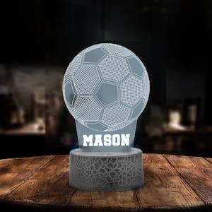 May include: A white 3D printed night light shaped like a soccer ball with the name "MASON" printed below. The night light is sitting on a wooden table.