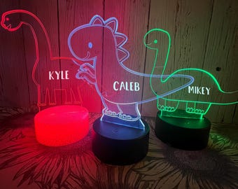 Puede incluir: Tres luces nocturnas de dinosaurio con bases LED que cambian de color. Los dinosaurios están iluminados en rojo, azul y verde. Los nombres "KYLE", "CALEB" y "MIKEY" están impresos en los dinosaurios.