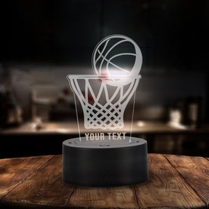 May include: A white 3D printed basketball and hoop night light with a black base. The base has the text "YOUR TEXT" on it.