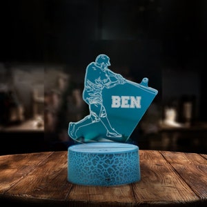 May include: A blue 3D printed night light with a baseball player silhouette and the name "BEN" on the base. The light is on a wooden table.