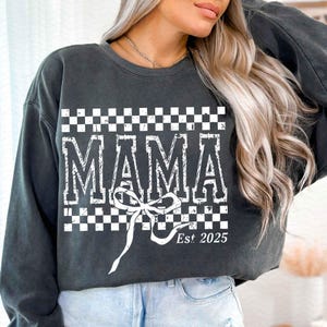 May include: A charcoal gray sweatshirt with a white checkered pattern and the word "MAMA" in a vintage font. The sweatshirt also features a white bow and the text "Est. 2025".