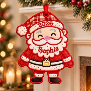 May include: A fabric Santa Claus Christmas ornament featuring a red and white plaid hat with the year 2026, a white beard, and a red suit with a black belt. The name Sophia is embroidered in red script across the center of the white beard.