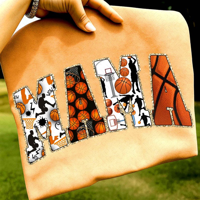 Puede incluir: Un art&iacute;culo de tela naranja con la palabra "MAMA" en letras grandes. Las letras est&aacute;n decoradas con balones de baloncesto, aros y siluetas de jugadores. Los bordes de las letras tienen un ribete dorado brillante.
