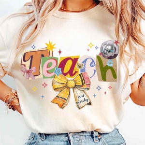 Op de afbeelding: Crèmekleurig T-shirt met het woord "Teach" in kleurrijke blokletters. Een grote, decoratieve strik en een discobal zijn in het ontwerp verwerkt. De strik heeft een label met "Mrs. Emily".