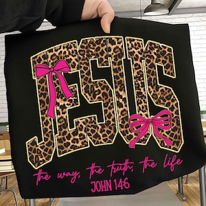 May include: Black t-shirt with the word "JESUS" in leopard print lettering, outlined in gold. Pink bows accent the letters. Below, the text reads "the way, the truth, the life" and "JOHN 14:6" in pink.