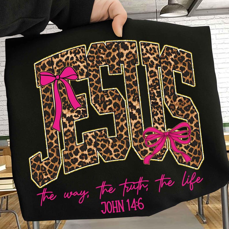 May include: Black t-shirt with the word "JESUS" in leopard print lettering, outlined in gold. Pink bows accent the letters. Below, the text reads "the way, the truth, the life" and "JOHN 14:6" in pink.