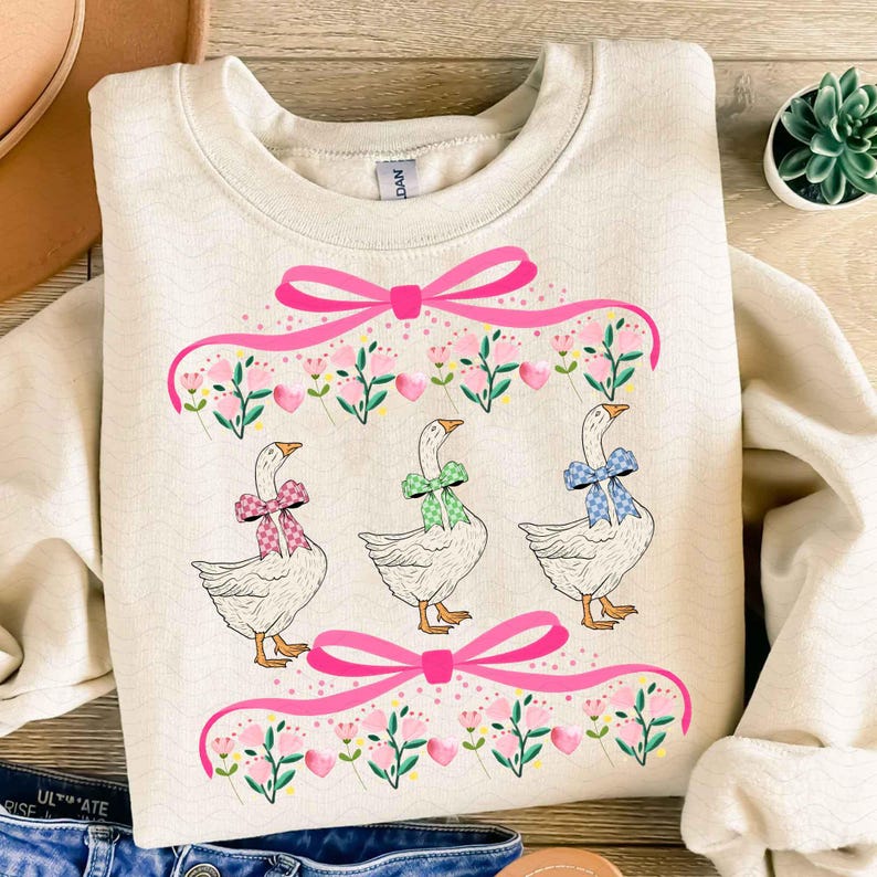 May include: Cream-colored sweatshirt featuring three geese with patterned bows. The geese are beneath a pink ribbon and floral design. The sweatshirt has a crew neck and long sleeves. The design includes pink bows and floral accents.