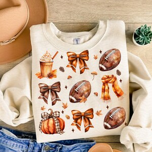 May include: Cream-colored sweatshirt with a fall-themed design. The design includes watercolor illustrations of footballs, bows, a pumpkin, a scarf, a coffee drink, and autumn leaves. The sweatshirt is made of a soft material.