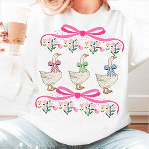 May include: White sweatshirt featuring three geese with colorful bowties. Pink bows and floral accents frame the geese. The geese have orange beaks and feet. The design is a whimsical, springtime theme.