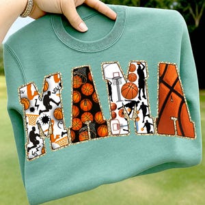 Puede incluir: Una sudadera verde azulado con la palabra "MAMA" en letras grandes y decorativas. Las letras est&aacute;n rellenas de balones de baloncesto, siluetas de baloncesto y redes. Las letras est&aacute;n delineadas con purpurina dorada.