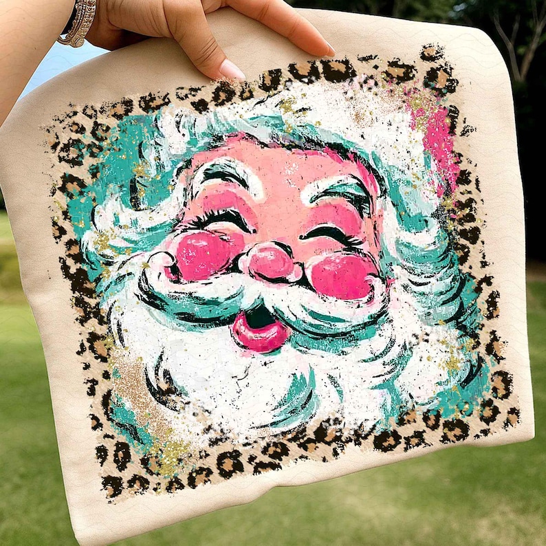 May include: A beige sweatshirt featuring a colorful Santa Claus face. The Santa has pink cheeks and lips, white beard and hair, and turquoise accents. A leopard print border surrounds the image, with gold glitter accents.