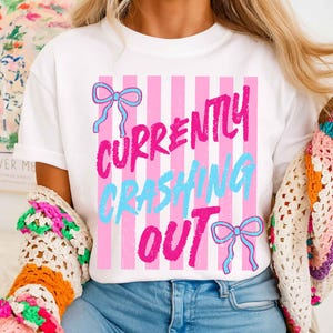 May include: White t-shirt with a pink and white striped design. The shirt features the text "CURRENTLY CRASHING OUT" in pink and blue, with two blue bow graphics. A casual, everyday tee.