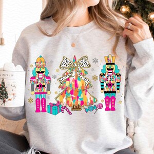 Puede incluir: Una sudadera gris con un árbol de Navidad colorido y dos figuras de cascanueces. El árbol está decorado con un lazo rosa y blanco y tiene un diseño de purpurina dorada. Los cascanueces llevan atuendos azules y rojos con detalles dorados.
