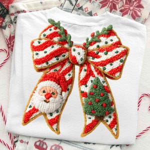 Pode incluir: T-shirt branco com tema natalino com um design de la&ccedil;o grande e detalhado. O la&ccedil;o &eacute; listrado em vermelho e branco com detalhes verdes e inclui um rosto do Papai Noel e uma &aacute;rvore de Natal. A camiseta est&aacute; dobrada em uma superf&iacute;cie branca.