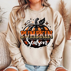May include: A beige sweatshirt with a graphic of a pumpkin with black and orange stripes and the text "Pumpkin Season" in black and orange glitter.