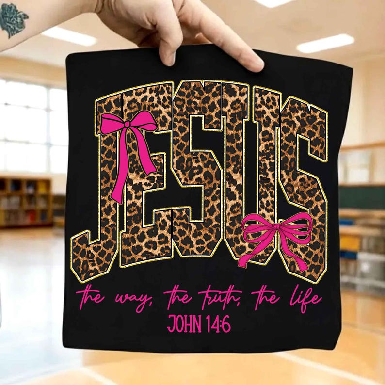 May include: Black t-shirt with the word "JESUS" in leopard print with gold trim and pink bows. The text "the way, the truth, the life" and "JOHN 14:6" are in pink script below the word "JESUS".