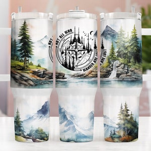 May include: A white stainless steel travel mug with a black and white compass design and the text "Not all who wander are lost". The mug features a watercolor design of mountains, trees, and a waterfall.