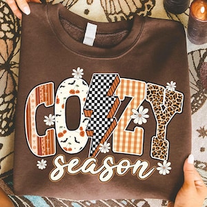 以下が含まれることがあります： 茶色のスウェットシャツ。装飾的なフォントで「COZY season」の文字がプリントされています。文字は、市松模様、チェック柄、花柄などの様々なパターンで埋められています。白いデイジーやその他の装飾要素がテキストを囲んでいます。