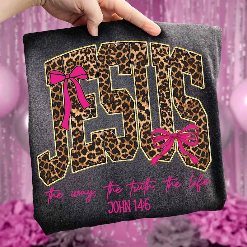 May include: A black t-shirt featuring the word "JESUS" in a leopard print design with gold trim and pink bows. The text "the way, the truth, the life" and "JOHN 14:6" are printed in pink below the word "JESUS".