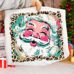May include: A white pillow with a colorful Santa Claus face design. The Santa face features pink cheeks and lips, a white beard, and a turquoise and white hat. The pillow has a leopard print border and gold glitter accents.