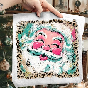 May include: A white tote bag featuring a colorful Santa Claus face with a pink nose and lips, and a white beard. The Santa face is surrounded by a leopard print border. The Santa's face has turquoise and gold accents.