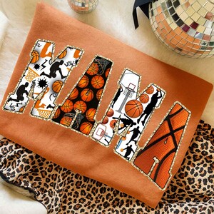 Puede incluir: Una sudadera naranja con la palabra "MAMA" en letras grandes con bordes brillantes. Cada letra est&aacute; decorada con dise&ntilde;os de baloncesto, incluyendo balones, aros y siluetas de jugadores. La sudadera est&aacute; sobre una tela con estampado de leopardo.