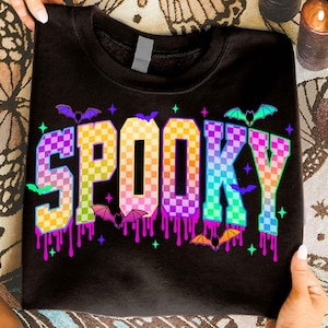 Könnte beinhalten: Schwarzes Sweatshirt mit dem Wort "SPOOKY" in einem karierten, regenbogenfarbenen Schriftzug. Das Design umfasst Fledermäuse und Sterne mit tropfenden Farbeffekten, ideal für Halloween.