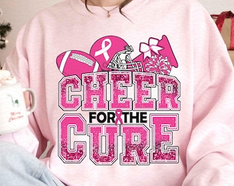 Op de afbeelding: Roze sweatshirt met een roze lint, een voetbal, cheerleader pompoenen en een voetbalhelm. De tekst luidt "Cheer for the Cure".