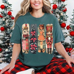 May include: Teal blue t-shirt featuring a vintage Christmas illustration of a nutcracker, a girl in a red dress, a Christmas tree, and a squirrel wearing a crown.