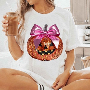Może przedstawiać: Biała koszulka z cekinową dynią z różową kokardką. Dynia ma twarz jack-o'-lantern.