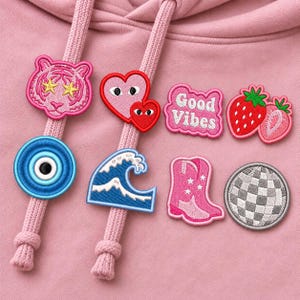 May include: A light pink hoodie featuring various embroidered patches including a pink tiger, a blue evil eye, a heart, a wave, a cowboy boot, a disco ball, a strawberry, and a pink speech bubble with the text Good Vibes.