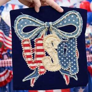 May include: A navy blue square with a patriotic design. The word "USA" is spelled out with red and white striped fabric for the "U", cream-colored fabric for the "S", and blue denim with white stars for the "A". A denim bow and pearl accents complete the design.