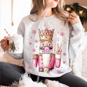 May include: A gray sweatshirt with a pink and gold watercolor illustration of a nutcracker holding a peace sign. The nutcracker is wearing a crown and has a pink bubble gum in its mouth.