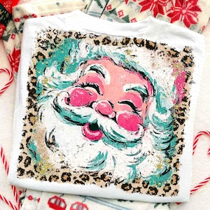 May include: White t-shirt featuring a Santa Claus graphic. The Santa face is pink with teal accents and a white beard. The design is framed by a leopard print border with gold glitter accents. Christmas themed apparel.
