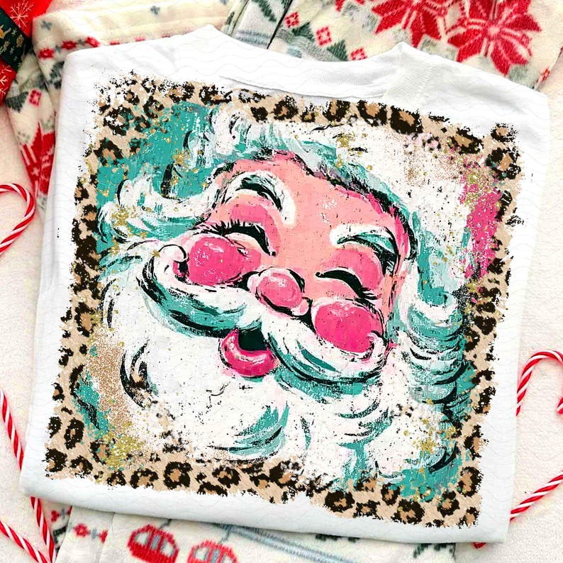 May include: White t-shirt featuring a Santa Claus graphic. The Santa face is pink with teal accents and a white beard. The design is framed by a leopard print border with gold glitter accents. Christmas themed apparel.