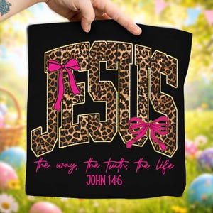 May include: Black tote bag with the word "JESUS" in large, leopard print letters outlined in gold. Pink bows accent the letters. The text "the way, the truth, the life" and "JOHN 14:6" are in pink script below the word "JESUS".