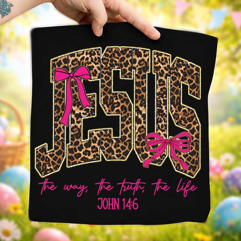 May include: Black tote bag with the word "JESUS" in large, leopard print letters outlined in gold. Pink bows accent the letters. The text "the way, the truth, the life" and "JOHN 14:6" are in pink script below the word "JESUS".