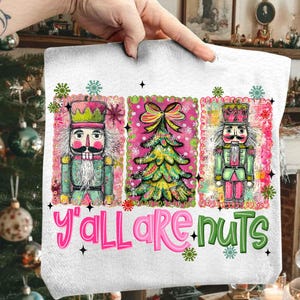 May include: White Christmas-themed towel with three panels. The panels feature a nutcracker, a Christmas tree, and another nutcracker. The text "Y'all are nuts" is written in pink and green. The towel is held by a hand.