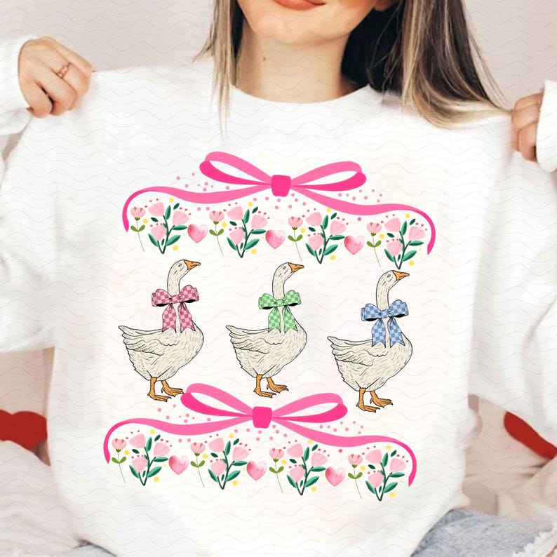 May include: White sweatshirt featuring a whimsical design of three geese with patterned bows. The geese are surrounded by pink bows, flowers, and hearts. The design is framed by a pink ribbon border.