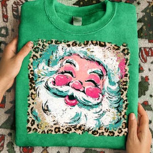 May include: Green sweatshirt featuring a colorful Santa Claus graphic. The Santa design has a pink face, white beard, and a leopard print border. The sweatshirt is folded and held by hands.