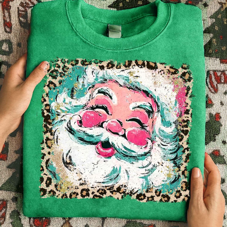 May include: Green sweatshirt featuring a colorful Santa Claus graphic. The Santa design has a pink face, white beard, and a leopard print border. The sweatshirt is folded and held by hands.