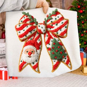 Pode incluir: Um la&ccedil;o de Natal festivo com sec&ccedil;&otilde;es listradas em vermelho e branco, um rosto de Papai Noel e uma &aacute;rvore de Natal verde. O la&ccedil;o &eacute; feito de um material texturizado com uma borda dourada e detalhes verdes. O la&ccedil;o &eacute; segurado por uma pessoa.
