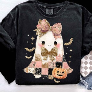 May include: Black sweatshirt featuring a cute ghost design with a pink bow, gold accents, and a pumpkin. The ghost has a checkered dress in pink, gold, and black. Halloween-themed apparel.