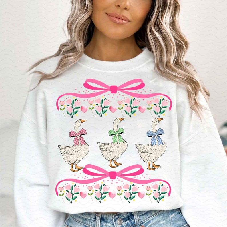May include: White sweatshirt featuring three geese with patterned bows. The geese are positioned below a pink ribbon and floral design. The sweatshirt has a crew neck and long sleeves.