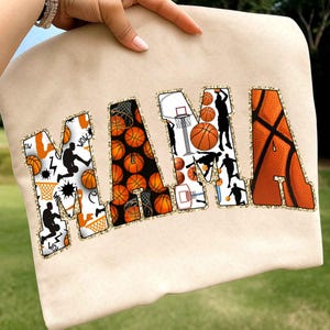 Puede incluir: Sudadera beige con la palabra "MAMA" en dise&ntilde;os de baloncesto naranja, negro y blanco. Las letras est&aacute;n delineadas con purpurina dorada. La "M" presenta siluetas de baloncesto y gr&aacute;ficos estilo c&oacute;mic.