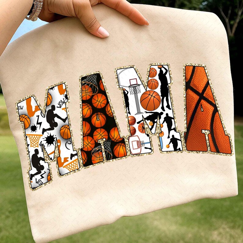 Puede incluir: Sudadera beige con la palabra "MAMA" en dise&ntilde;os de baloncesto naranja, negro y blanco. Las letras est&aacute;n delineadas con purpurina dorada. La "M" presenta siluetas de baloncesto y gr&aacute;ficos estilo c&oacute;mic.