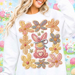 Puede incluir: Sudadera blanca con un diseño colorido de Pascua. El diseño incluye un conejito rodeado de flores en tonos rosas, amarillos y azules. La sudadera tiene cuello redondo y mangas largas, ideal para la primavera.