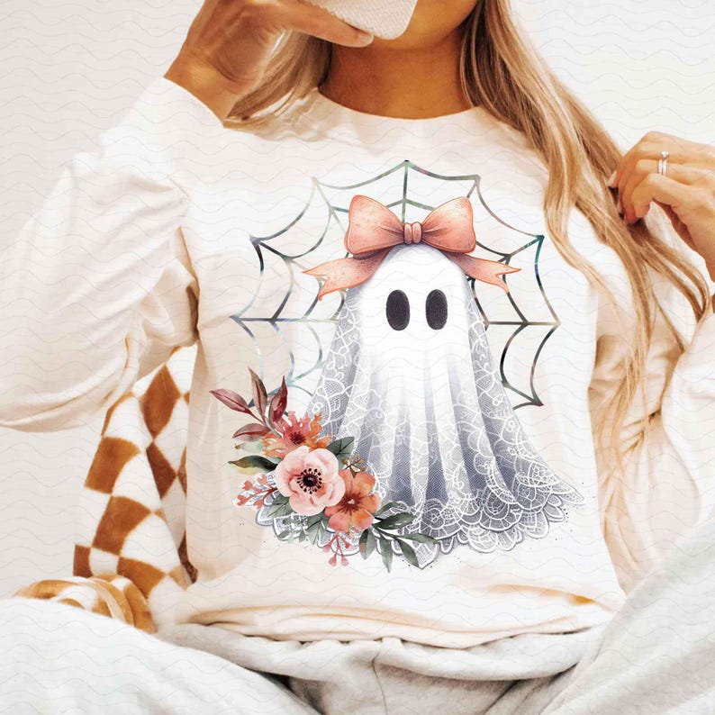 May include: A white long-sleeved shirt with a graphic of a ghost wearing a pink bow and surrounded by a spiderweb. The ghost is wearing a lace overlay and there are floral accents around the bottom of the graphic.